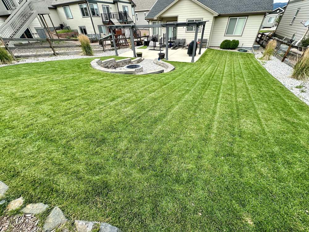 A mowed backyard with a stone fire pit, pergola, and planting beds along the fence