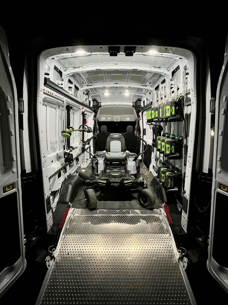 Inside the van: a battery powered ride-on mower on a loading ramp, with trimmers and a wall of charged batteries racked along one side